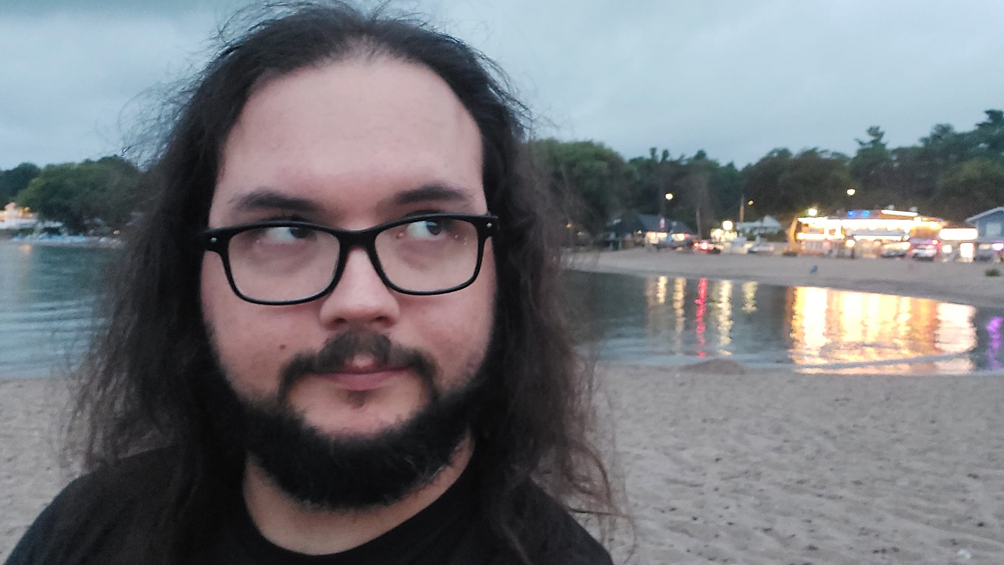 A man with long dark hair and a beard is at a sandy beach with a granite boulder breakwater, photo 4